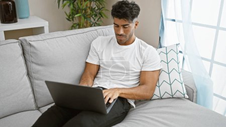 Handsome hispanic man using a laptop while sitting comfortably on a couch in a well-lit living room.