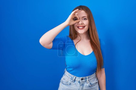 Téléchargez les photos : Femme rousse debout sur fond bleu faisant un geste correct avec la main souriante, les yeux regardant à travers les doigts avec un visage heureux. - en image libre de droit