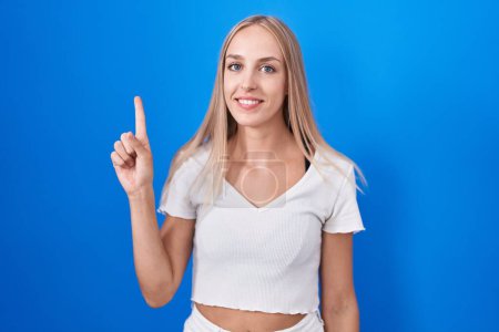 Photo for Young caucasian woman standing over blue background showing and pointing up with finger number one while smiling confident and happy. - Royalty Free Image