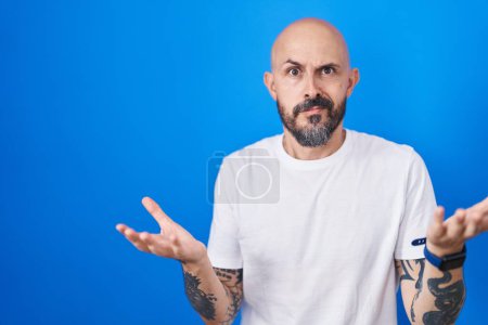 Photo for Hispanic man with tattoos standing over blue background clueless and confused with open arms, no idea concept. - Royalty Free Image