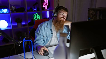 Foto de Aburrido joven pelirrojo streamer conseguir cansado, luchando para mantenerse despierto jugando videojuego, envuelto en la noche oscura en su sala de juegos - Imagen libre de derechos