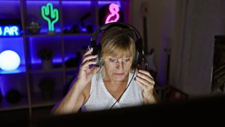 Schöne blonde Frau mittleren Alters Streamer ernsthaft eingetaucht, trägt Kopfhörer, Videospiel. Indoor-Nacht-Gaming-Raum Streaming mit fortschrittlichem Tech-Gadget.