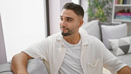 Foto de Hombre guapo con barba en una moderna sala de estar mirando hacia otro lado pensativamente. - Imagen libre de derechos