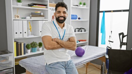 Téléchargez les photos : Jeune homme hispanique avec la barbe souriant en toute confiance tout en se tenant les bras croisés dans une clinique de réadaptation moderne intérieur - en image libre de droit
