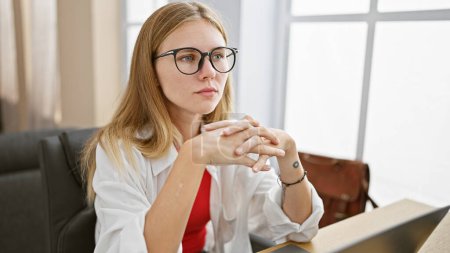 Téléchargez les photos : Une jeune femme réfléchie avec des lunettes et des cheveux blonds contemplant dans un bureau. - en image libre de droit