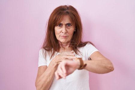 Foto de Mujer de mediana edad de pie sobre fondo rosa con prisa apuntando a ver el tiempo, la impaciencia, molesto y enojado por retraso plazo - Imagen libre de derechos