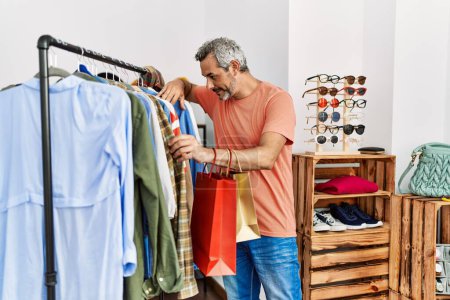 Téléchargez les photos : Moyen âge homme aux cheveux gris client souriant confiant tenant des vêtements de rack au magasin de vêtements - en image libre de droit