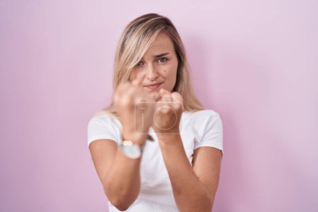 Téléchargez les photos : Jeune femme blonde debout sur fond rose prêt à se battre avec geste de défense poing, visage en colère et bouleversé, peur du problème - en image libre de droit