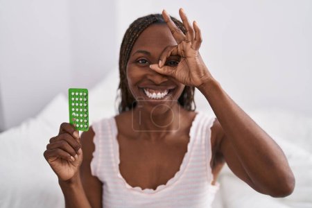 Téléchargez les photos : Femme afro-américaine tenant des pilules contraceptives souriant heureux faire ok signe avec la main sur les yeux regardant à travers les doigts - en image libre de droit