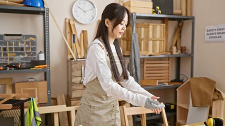 Photo for A young asian woman in a carpentry workshop wearing gloves and an apron stands surrounded by wood and tools. - Royalty Free Image