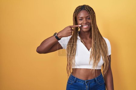 Téléchargez les photos : Femme afro-américaine avec les cheveux tressés debout sur fond jaune pointant avec le doigt de la main sur le visage et le nez, souriant gai. concept de beauté - en image libre de droit