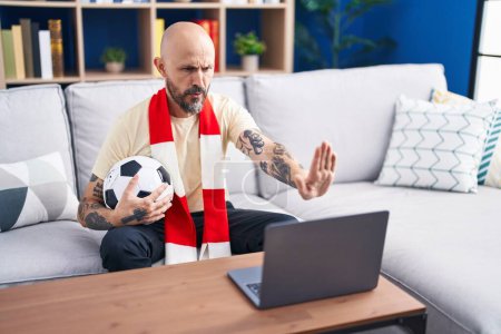 Téléchargez les photos : Homme hispanique avec tatouages regarder match de football hooligan tenant ballon sur l'ordinateur portable avec la main ouverte faire stop signe avec expression sérieuse et confiante, geste de défense - en image libre de droit