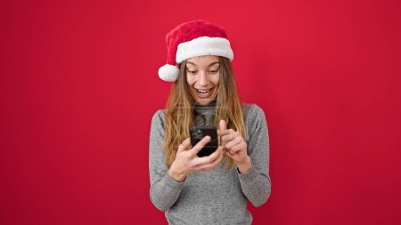 Téléchargez les photos : Jeune femme caucasienne portant un chapeau de Noël à l'aide d'un smartphone avec expression surprise sur fond rouge isolé - en image libre de droit