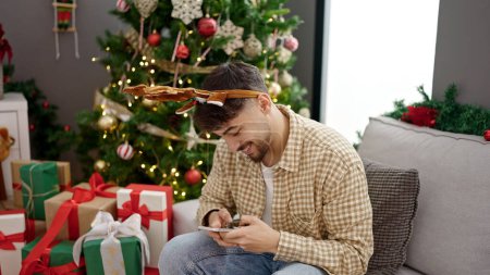 Foto de Hombre árabe joven usando teléfono inteligente sentado junto al árbol de Navidad en casa - Imagen libre de derechos