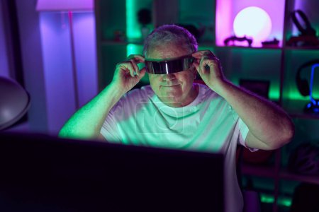 Téléchargez les photos : Homme aux cheveux gris moyen âge streamer jouer à un jeu vidéo en utilisant des lunettes de réalité virtuelle à la salle de jeux - en image libre de droit