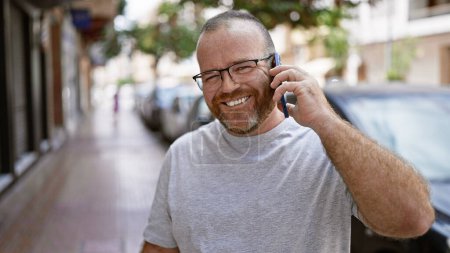 Hombre caucásico guapo y barbudo charlando con confianza en su teléfono inteligente, absorto positivamente en la divertida y soleada calle de la ciudad al aire libre, retratando un estilo de vida informal y fresco con una sonrisa cálida