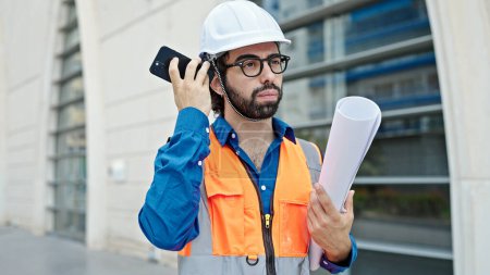 Photo for Young hispanic man architect holding blueprints listening to voice message by smartphone at construction place - Royalty Free Image