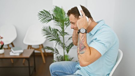 Photo for Hispanic man with a beard and tattoo, wearing headphones, sitting in an indoor lobby with plants and white chairs. - Royalty Free Image