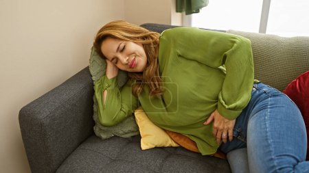 Foto de Una joven hispana sintiendo dolor de estómago mientras descansa en su casa en un sofá. - Imagen libre de derechos