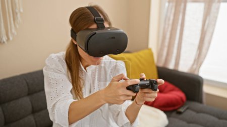 Foto de Mujer caucásica joven disfrutando de los auriculares de realidad virtual en casa, sentado en el sofá gris que sostiene el controlador de juego. - Imagen libre de derechos