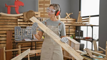Photo for A young woman wearing safety gear holds lumber in a well-equipped carpentry workshop - Royalty Free Image