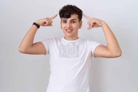 Téléchargez les photos : Jeune homme non binaire portant un t-shirt blanc décontracté souriant pointant vers la tête avec les deux doigts des mains, bonne idée ou pensée, bonne mémoire - en image libre de droit