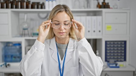 Foto de Una joven rubia con una bata de laboratorio ajusta sus gafas en un laboratorio - Imagen libre de derechos