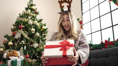 Téléchargez les photos : Jeune femme blonde déballer cadeau assis sur le canapé par arbre de Noël à la maison - en image libre de droit