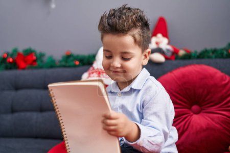Photo for Adorable hispanic toddler writing letter to santa claus sitting on sofa by christmas decoration at home - Royalty Free Image