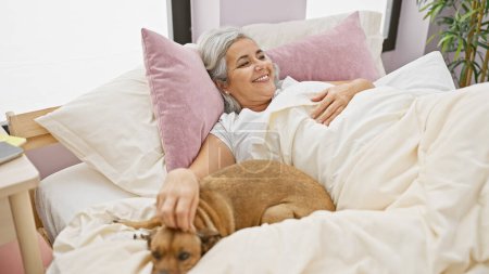 Foto de Una mujer sonriente de mediana edad disfruta de un momento de relax en la cama con su perro mascota, capturando una sensación de comodidad y compañía en el interior. - Imagen libre de derechos