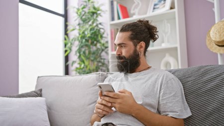 Téléchargez les photos : Un jeune homme barbu se prélasse sur un canapé gris à la maison, absorbé dans son smartphone, représentant la vie moderne décontractée. - en image libre de droit