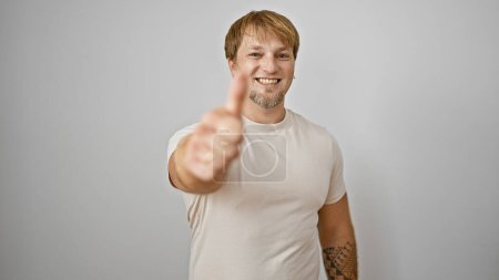 Un jeune homme joyeux avec une barbe et un tatouage donnant un pouce vers le haut sur un fond blanc.