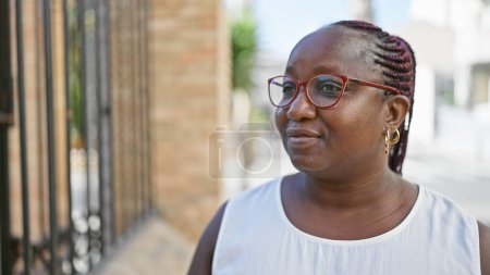 Foto de Cool mujer afroamericana de tamaño grande en gafas, con trenzas, lanzando una expresión seria mientras mira a un lado en una calle urbana soleada. - Imagen libre de derechos