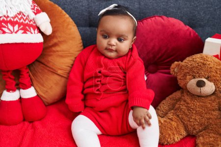 Téléchargez les photos : Africain américain bébé souriant confiant assis sur le canapé avec poupée santa claus à la maison - en image libre de droit