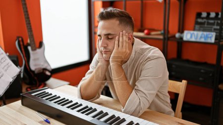 Téléchargez les photos : Un jeune homme hispanique cher assis au piano dans un studio de musique moderne, reflétant la créativité et la contemplation. - en image libre de droit