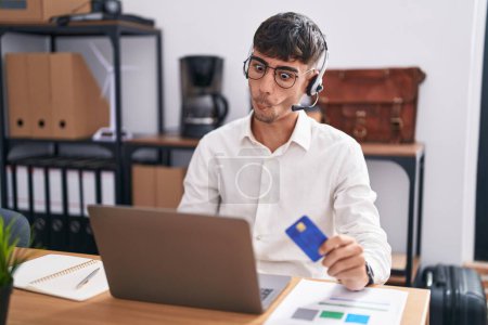Photo for Young hispanic man working using computer laptop holding credit card making fish face with lips, crazy and comical gesture. funny expression. - Royalty Free Image
