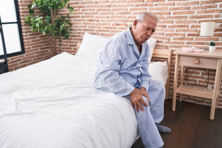 Foto de Hombre de pelo gris de mediana edad que sufre de lesión de rodilla sentado en la cama en el dormitorio - Imagen libre de derechos
