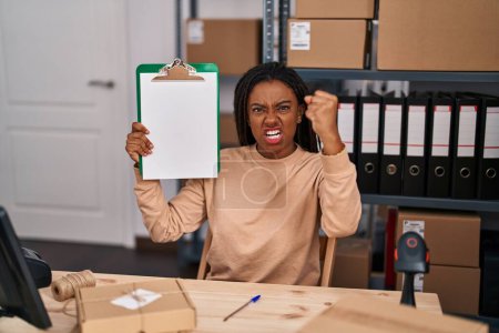 Téléchargez les photos : Jeune afro-américain avec des tresses travaillant dans une petite entreprise e-commerce montrant presse-papiers ennuyé et frustré criant de colère, criant fou de colère et la main levée - en image libre de droit
