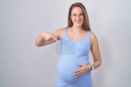 Foto de Mujer embarazada joven de pie sobre fondo blanco mirando confiado con sonrisa en la cara, señalándose con los dedos orgullosos y felices. - Imagen libre de derechos