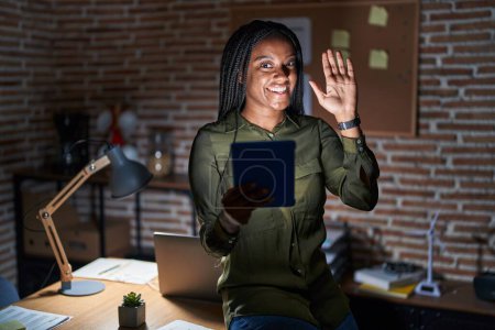 Téléchargez les photos : Jeune afro-américain avec des tresses travaillant au bureau la nuit renonçant à dire bonjour heureux et souriant, geste de bienvenue amical - en image libre de droit
