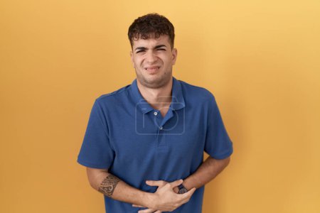 Téléchargez les photos : Jeune homme hispanique debout sur fond jaune avec la main sur le ventre parce que l'indigestion, la maladie douloureuse se sent mal. concept de douleur. - en image libre de droit