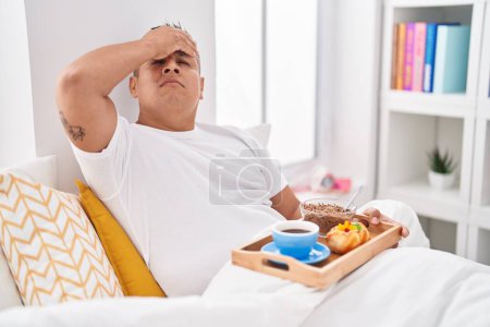 Téléchargez les photos : Jeune homme hispanique manger le petit déjeuner dans le lit stressé et frustré avec la main sur la tête, visage surpris et en colère - en image libre de droit