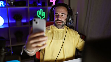 Un hombre barbudo sonriente con una sudadera amarilla se toma una selfie en una sala de juegos iluminada por neón por la noche.