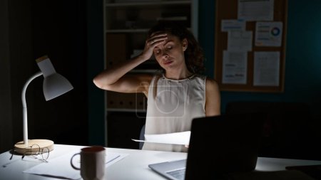 Photo for A stressed young woman works late at the office, illuminated by a desk lamp with a laptop, paperwork, and coffee mug. - Royalty Free Image