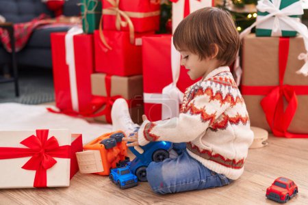 Foto de Adorable niño jugando con el juguete del camión sentado en el suelo por el árbol de Navidad en casa - Imagen libre de derechos