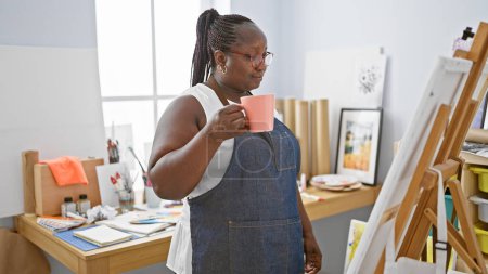 Téléchargez les photos : Femme artiste afro-américaine, debout avec une tasse de café, regardant son dessin dans son atelier d'art. - en image libre de droit