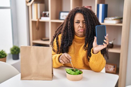 Photo for Plus size hispanic woman eating take away salad holding smartphone skeptic and nervous, frowning upset because of problem. negative person. - Royalty Free Image