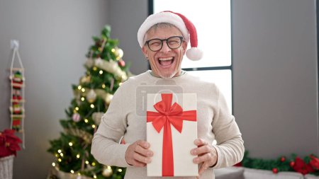 Photo for Middle age grey-haired man holding gift standing by christmas tree at home - Royalty Free Image
