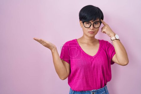 Photo for Young asian woman with short hair standing over pink background confused and annoyed with open palm showing copy space and pointing finger to forehead. think about it. - Royalty Free Image