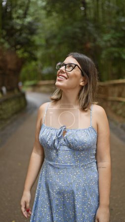 Téléchargez les photos : Belle femme hispanique avec des lunettes, souriant avec confiance alors qu'elle se tient parmi la forêt de bambous luxuriante de kyoto, regardant autour de elle, pleine de joie et d'étonnement. - en image libre de droit